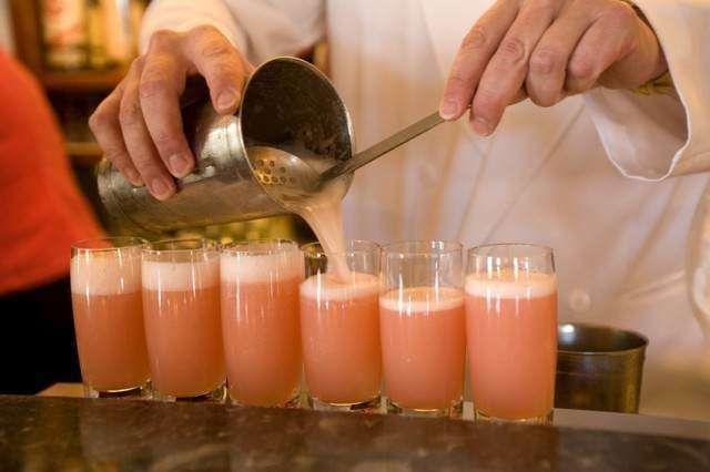 A bartender pours a pale pink cocktail from a stainless steel shaker into a row of glasses on a bar counter.
