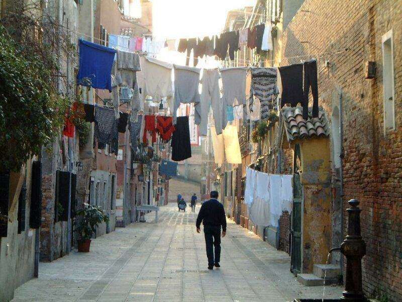A person walks down a narrow, sunlit stone alleyway in Italy with laundry hanging on lines stretched between buildings.