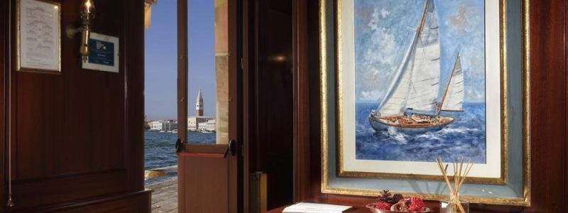 A view from a wooden-paneled room looking out at the Venice lagoon, featuring a framed sailboat painting on the wall.