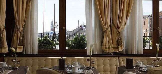 An elegant dining room with tan curtains and tufted booths overlooking a distant church tower through large windows.