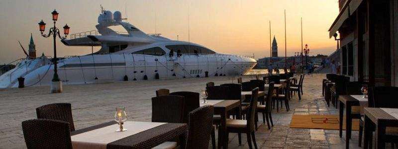 A waterfront restaurant patio at sunset with an empty table in the foreground and a large white yacht docked behind it.