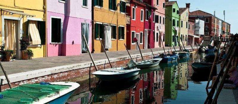 A row of brightly colored houses lines a canal in Burano, Italy, with small wooden boats docked along the water's edge.