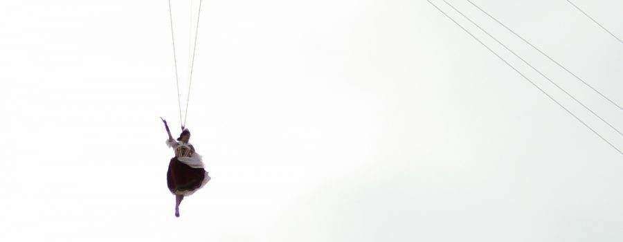 A person in a dark skirt and white top suspended in mid-air by cables against a bright, hazy sky.