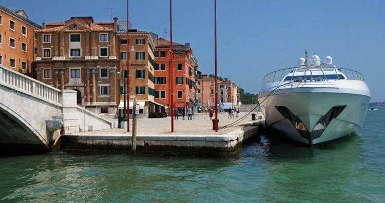 A large white yacht docked at a stone quay next to traditional orange buildings and a stone bridge in Venice.