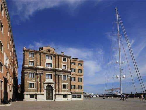 A historic multi-story building with a brown stone facade stands in a paved square beside a tall sailboat mast in Venice.