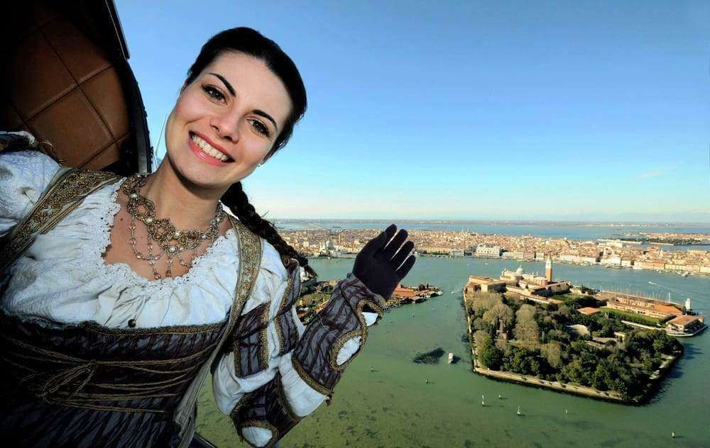 A smiling person in historical-style dress gestures toward an aerial view of San Giorgio Maggiore island in Venice.