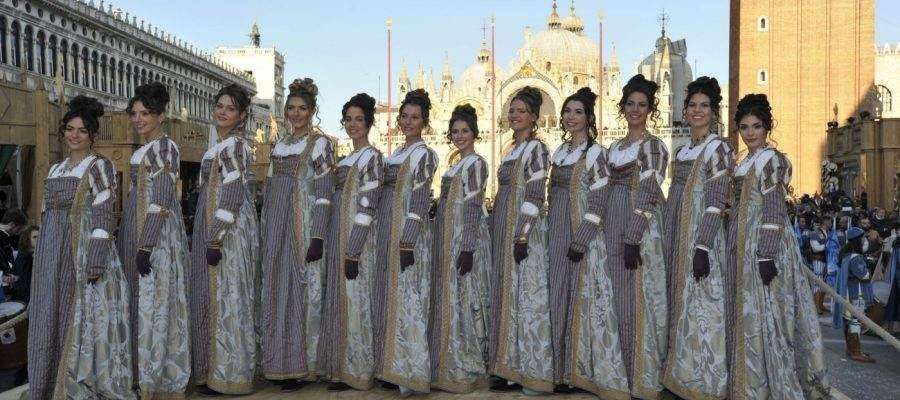A line of eleven people in identical historical-style gowns standing in St. Mark's Square, Venice, against a cathedral.