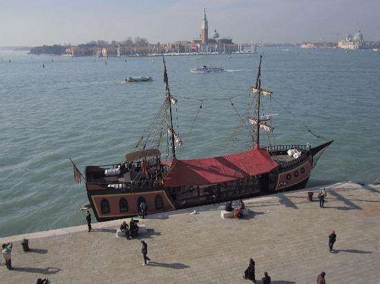 A tall, wooden sailing ship with red deck covers is docked at a stone quay in Venice, with people walking nearby.