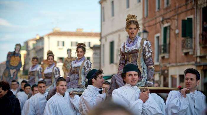 People in historical costume are carried through a street by individuals wearing white uniforms during an outdoor parade.