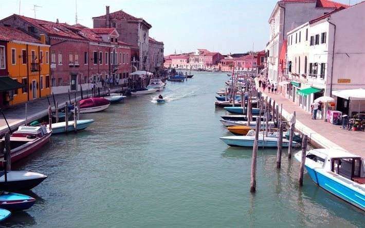 A canal in Murano, Italy, with colorful buildings on either side, moored boats, and a small boat traveling in the water.