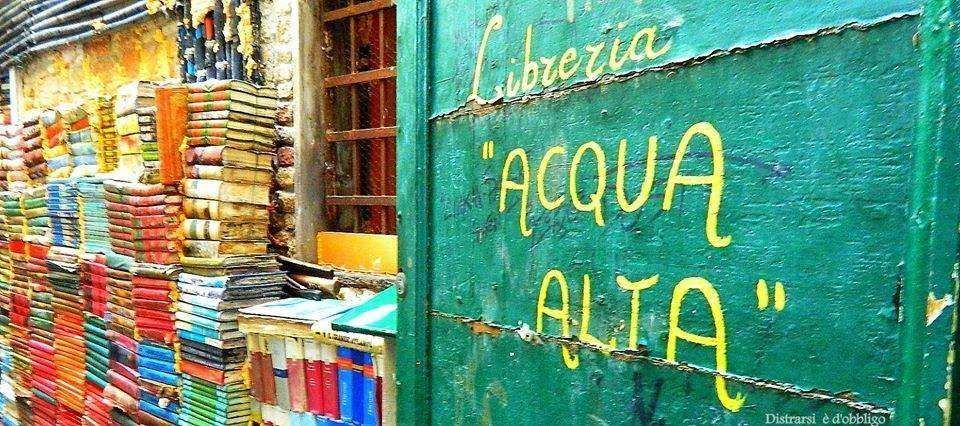 Stacks of colorful books inside the Libreria Acqua Alta in Venice, with the store name painted in yellow on a green door.