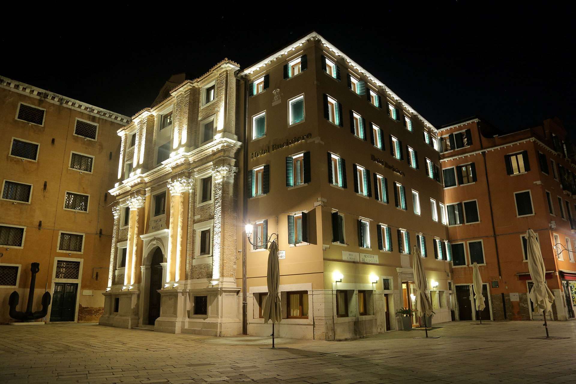A night shot of an ornate, warmly lit building complex in Venice, Italy, facing a paved square.