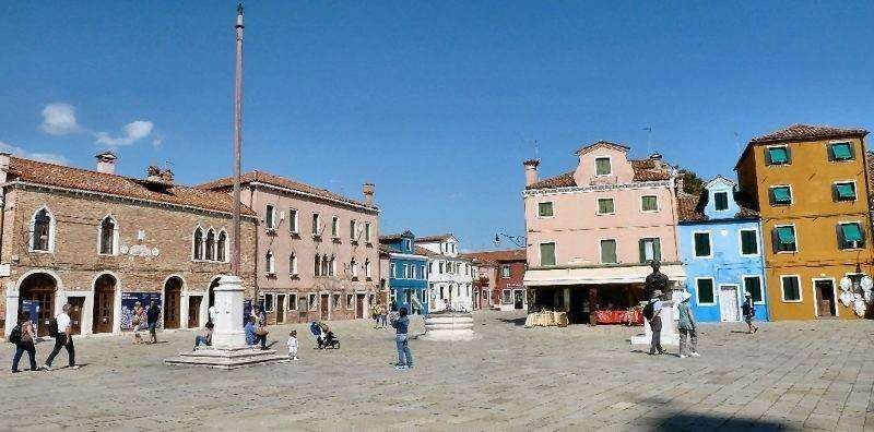 People walk through a sunny, paved town square in Burano, Italy, surrounded by vibrant, multi-colored historic buildings.