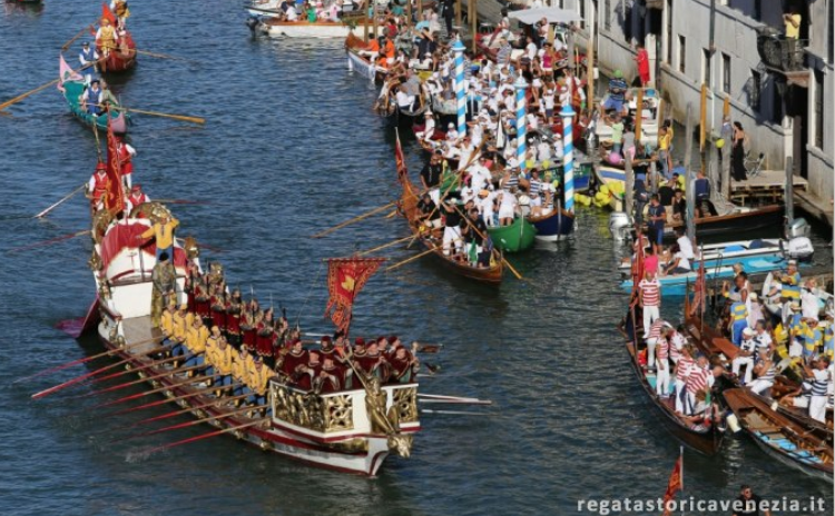 A decorated, multi-oared boat leads a colorful historical procession along a Venice canal, surrounded by smaller boats.