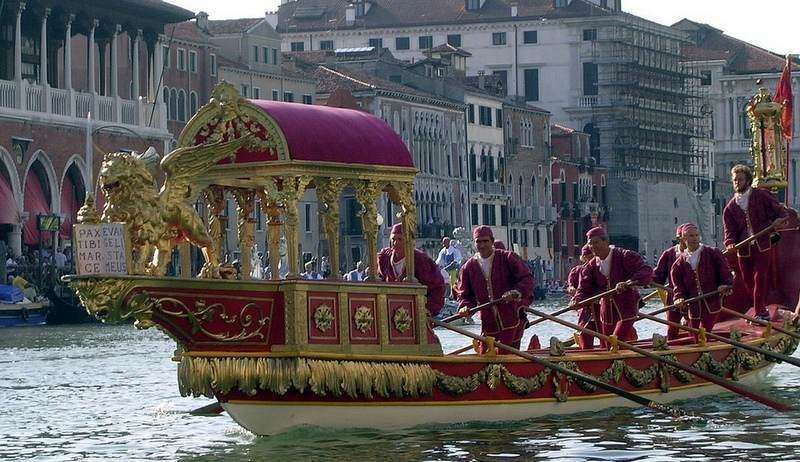 A traditional, ornate Venetian ceremonial boat decorated in red and gold floats on a canal, rowed by figures in red.