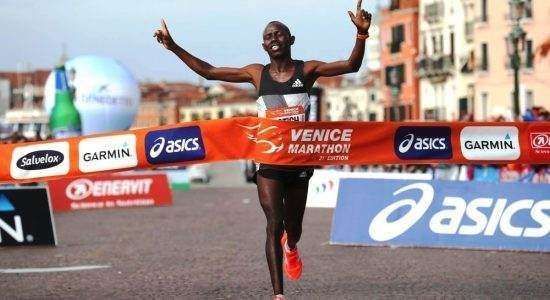 A runner celebrating with arms raised while crossing the finish line at the Venice Marathon under a bright banner.
