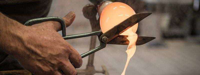 A artisan uses metal shears to trim molten, glowing orange glass while shaping it in a workshop.