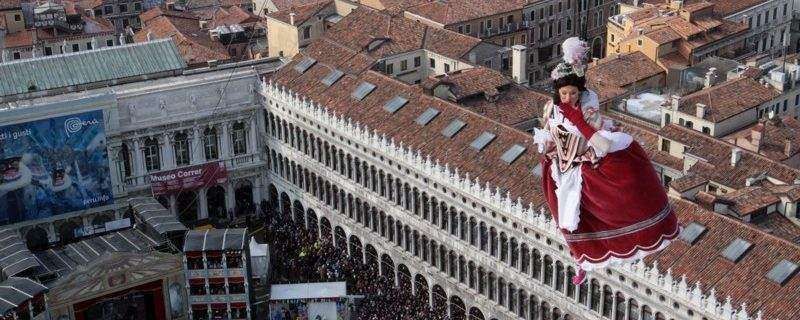 A costumed performer in a red dress suspended high above St. Mark's Square in Venice during the Carnival.