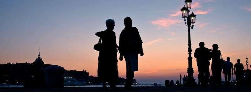 Silhouetted people walk along a waterfront at sunset near a decorative lamp post, with a distant cityscape in the background.