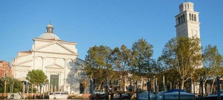 A white church with a dome and a tall bell tower standing beside a canal in Venice under a clear blue sky.