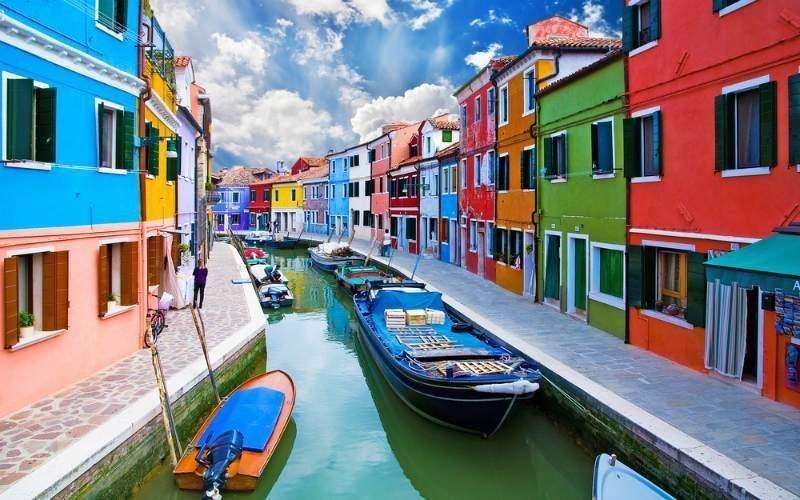 A canal in Burano, Italy, lined with vibrant, multicolored houses and moored boats under a blue, cloudy sky.