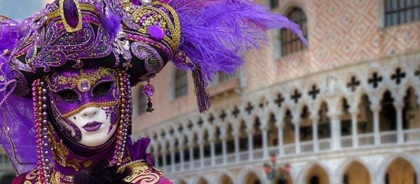 A person wearing an ornate purple Venetian carnival mask and feathered hat stands before the Doge’s Palace in Venice.