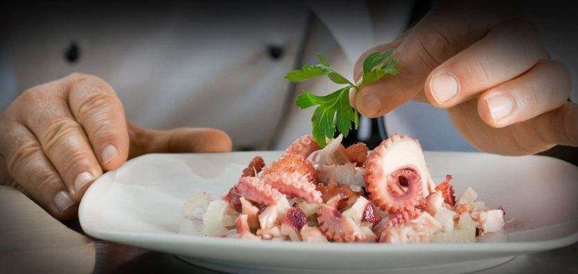 A chef in a white uniform garnishes a plate of chopped octopus salad with a fresh green parsley leaf.