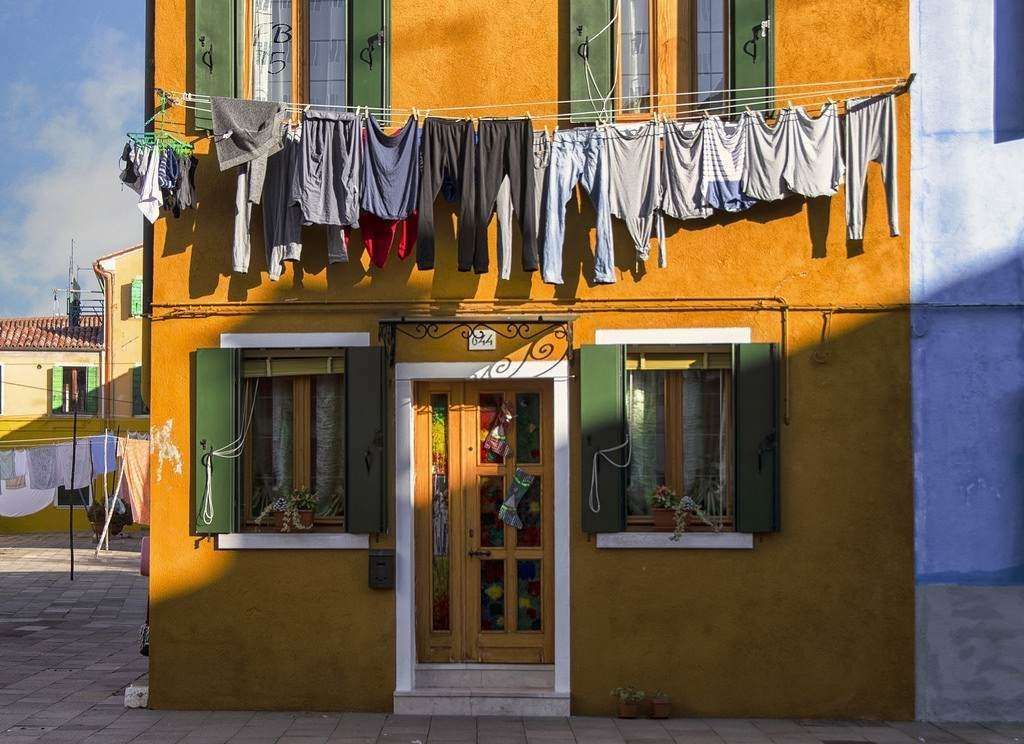 Laundry hangs from a line strung across the facade of a vibrant yellow house with green shutters in Burano, Italy.