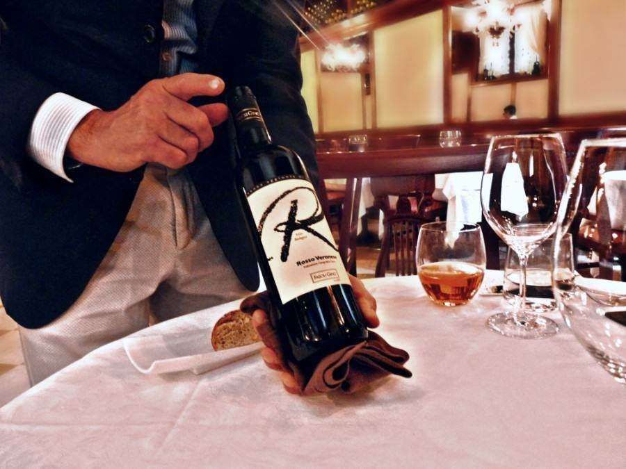 A server in a restaurant holds a bottle of red wine with a prominent 'R' on the label over a table set with glasses.