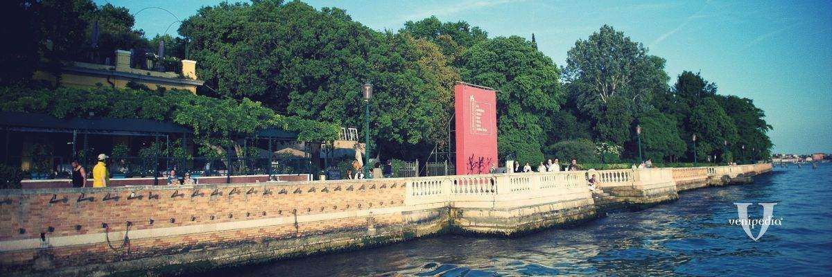 A waterfront park in Venice with a stone promenade, lush trees, and a prominent red monument along the lagoon.