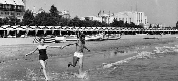 A couple runs hand-in-hand through the shallow water of a beach, with a long line of cabanas and buildings in the background.