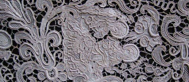 Intricate, cream-colored needle lace with swirling floral and organic motifs against a dark background.