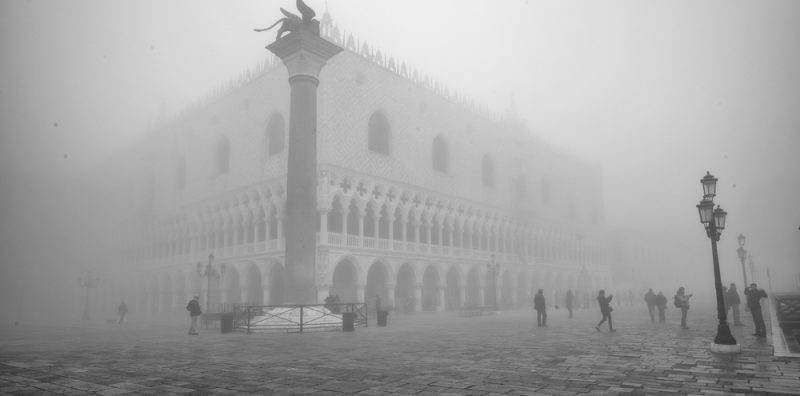 The Doge's Palace in Venice, Italy, partially obscured by thick fog, with pedestrians walking in the foreground.