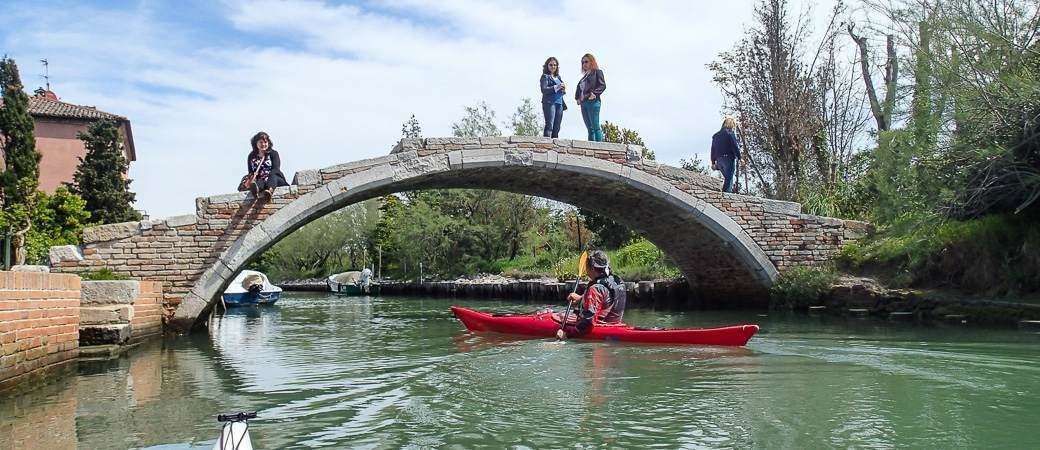 A red kayak floats under a stone bridge while several people stand on top of the bridge.
