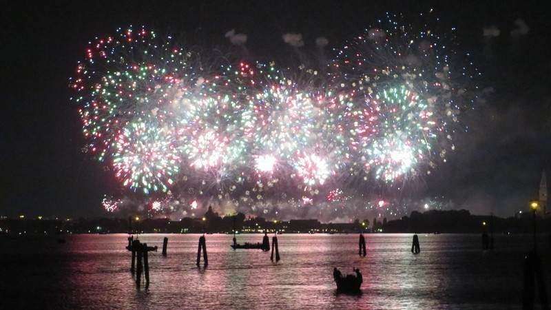 Fireworks displays in shades of white, pink, and green light up the night sky over a calm body of water with wooden pilings.