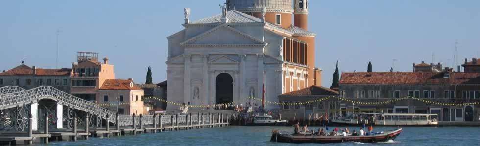 Venice’s San Giorgio Maggiore church stands by the water with a long wooden pedestrian bridge and a gondola in the harbor.
