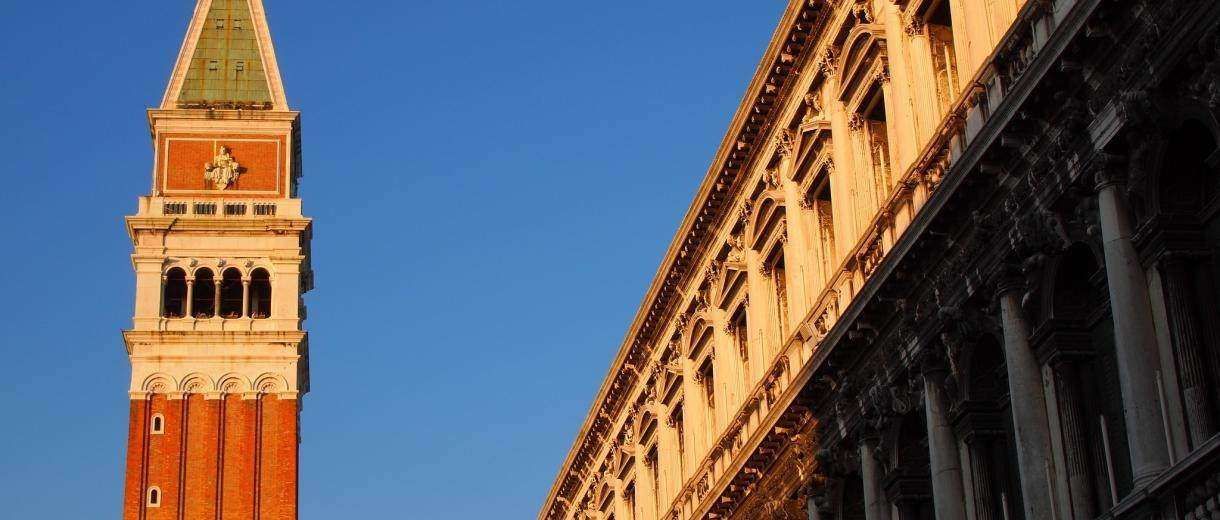 St. Mark's Campanile bell tower stands next to a sunlit, historic stone building with arched windows in Venice, Italy.