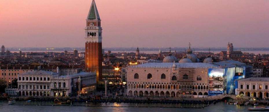Venice, Italy at sunset, featuring St. Mark's Campanile and the Doge's Palace illuminated against a pink sky.