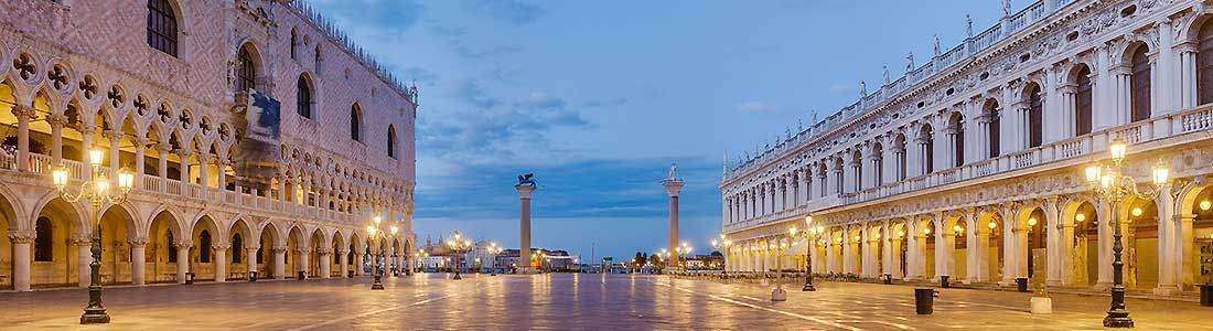 St. Mark's Square in Venice, Italy, showing the Doge's Palace and Procuratie Nuove at twilight with lit streetlamps.