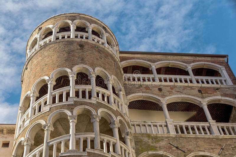 A stone cylindrical tower with spiraling open-arched balconies stands against a blue sky with soft, wispy clouds.