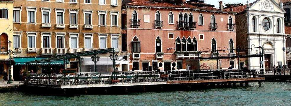 A waterfront patio with tables and chairs sits in front of historic buildings along a canal in Venice, Italy.