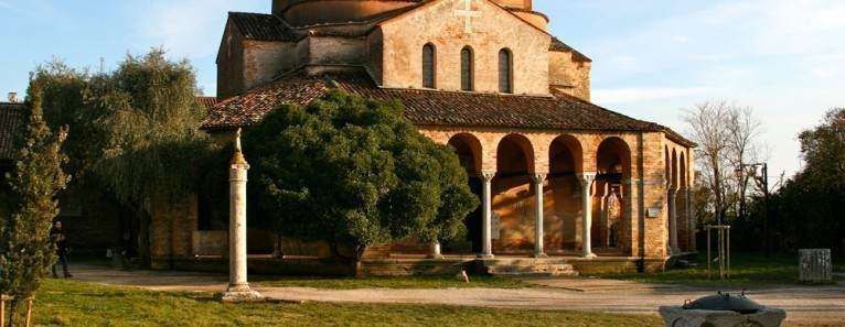 A tan stone church with a rounded portico and arched columns stands in a grassy courtyard at sunset.