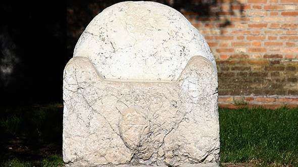 A rounded, weathered stone chair or throne sits outdoors against a brick wall and green grass.