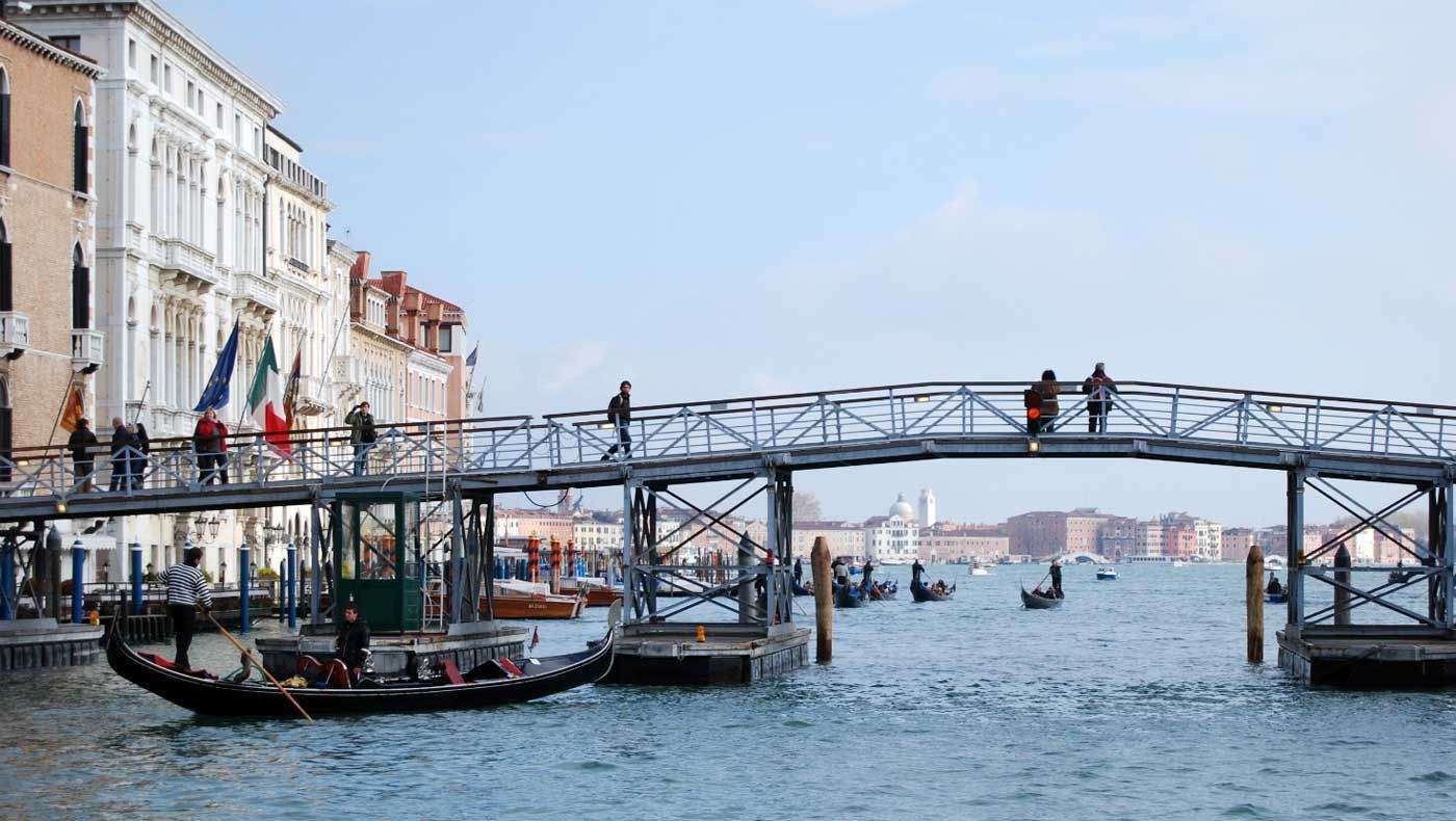 A gondola passes under a metal pedestrian bridge in Venice, with buildings along the canal and people walking above.