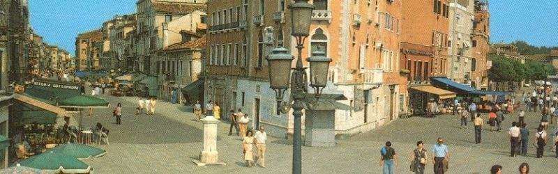 A lively European square with pedestrians, outdoor cafe seating, historic buildings, and an ornate street lamp.