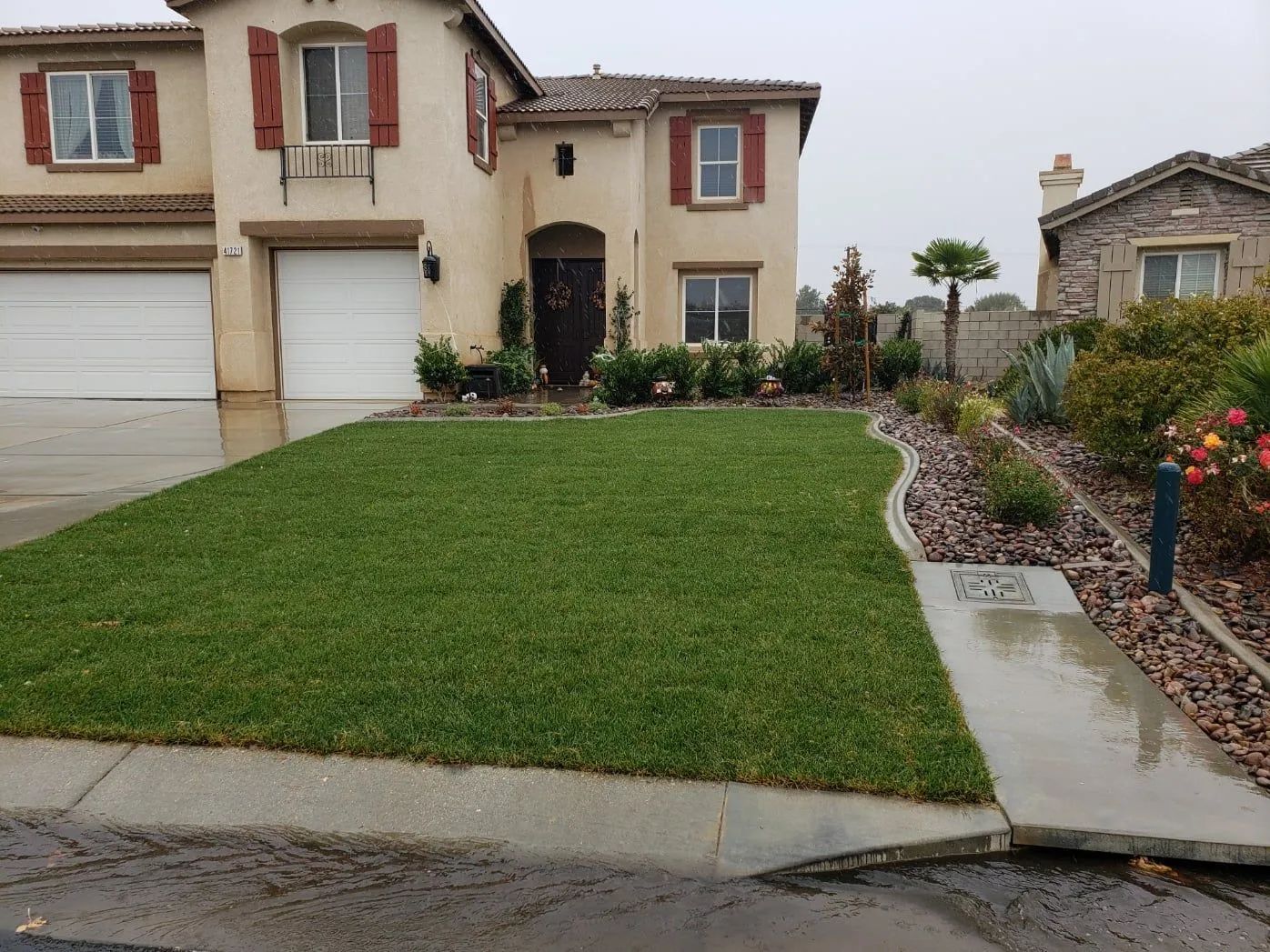 Two-story beige house with a green lawn, sidewalk, and landscaping on a rainy day.
