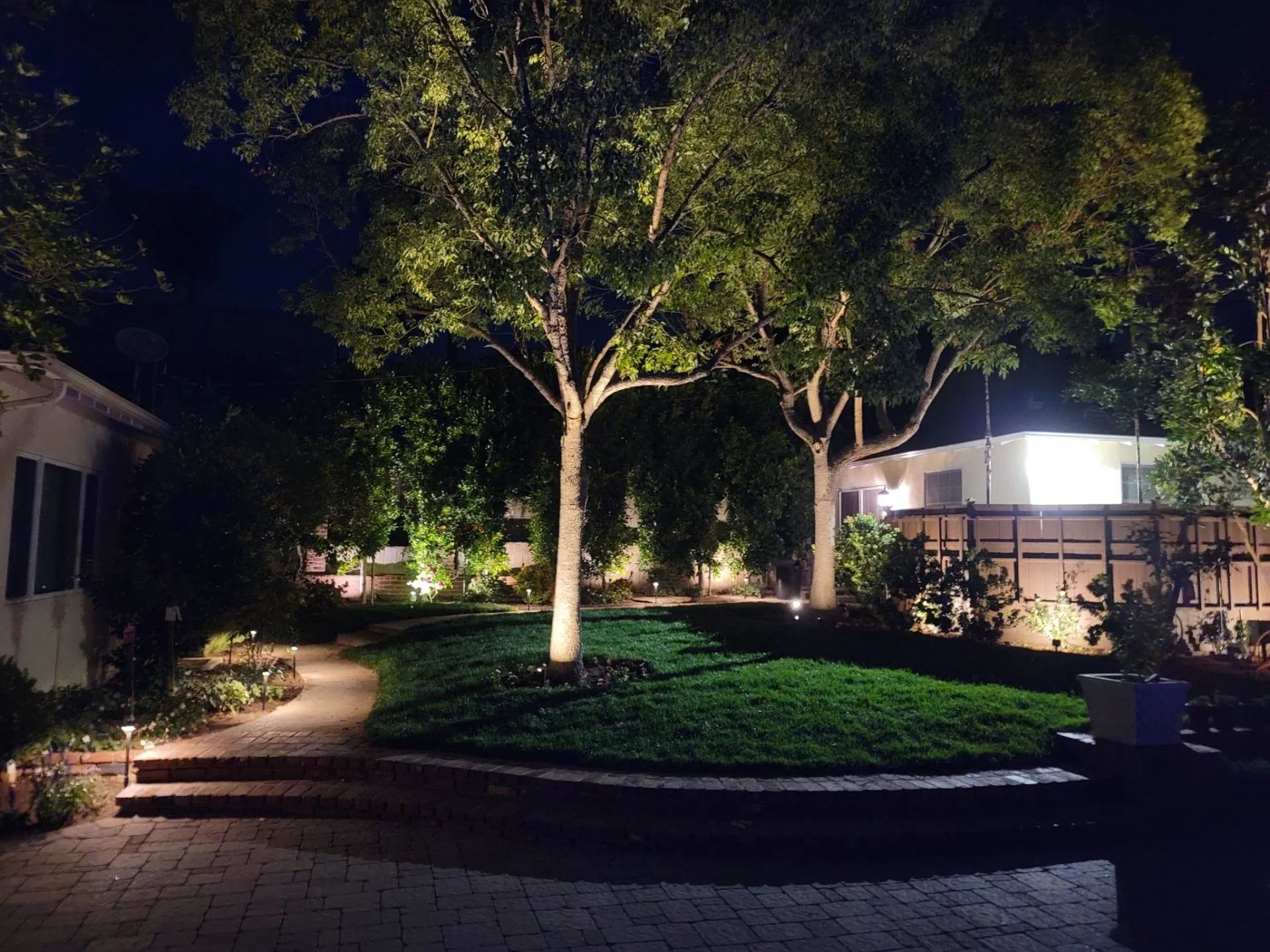 Night-lit garden with trees, brick pathway, and tiered lawn.
