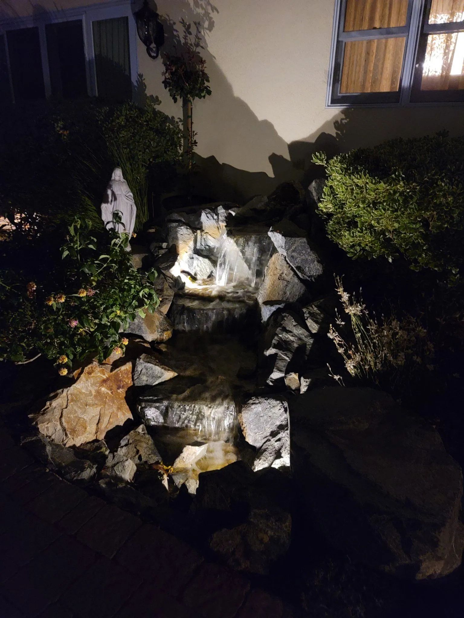 A tiered stone water fountain in a garden at night, illuminated by soft spotlights against the wall of a home.