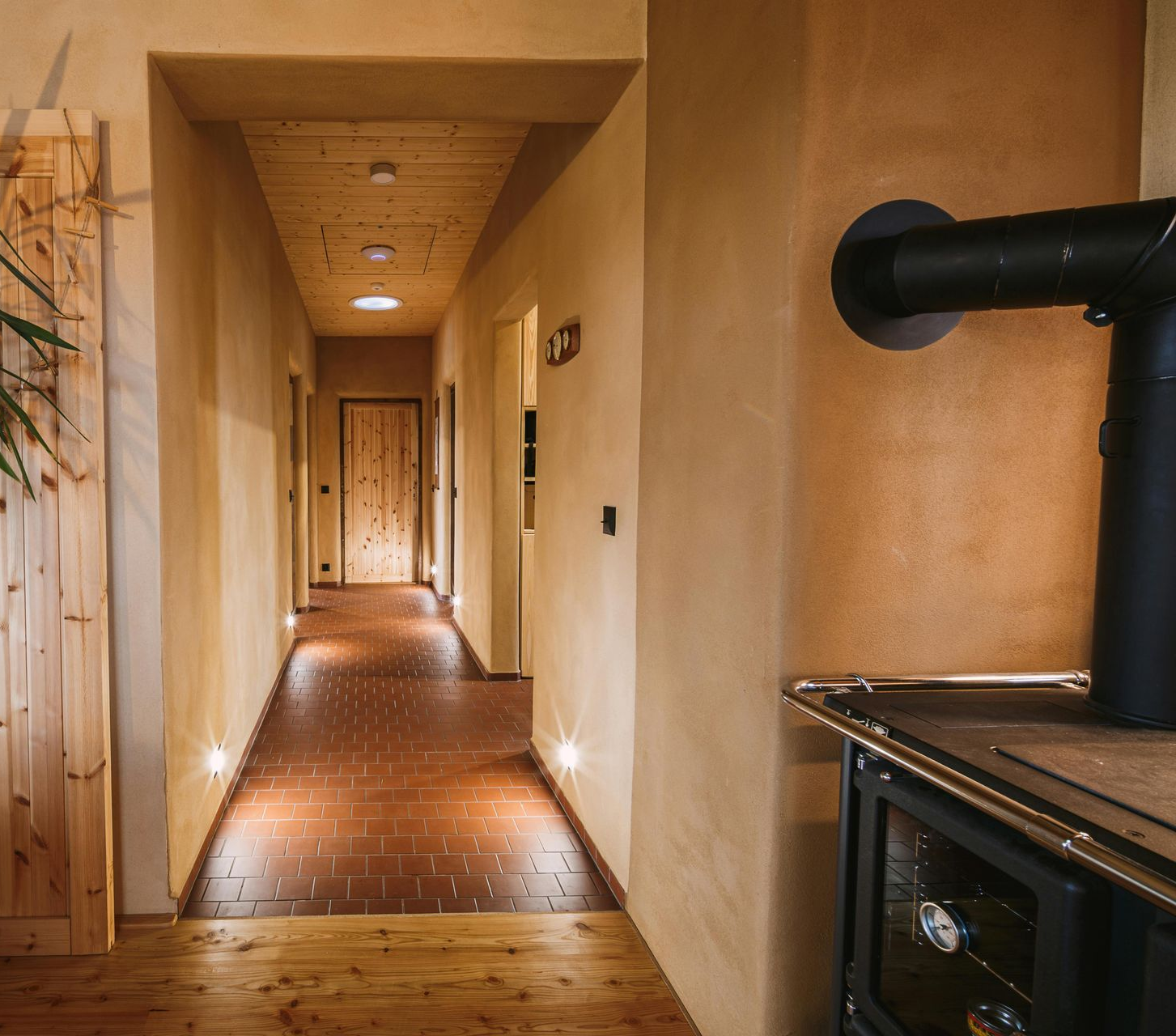 Long hallway with wooden ceiling and floor, illuminated by lights, leads to a door; a stove is on the right.