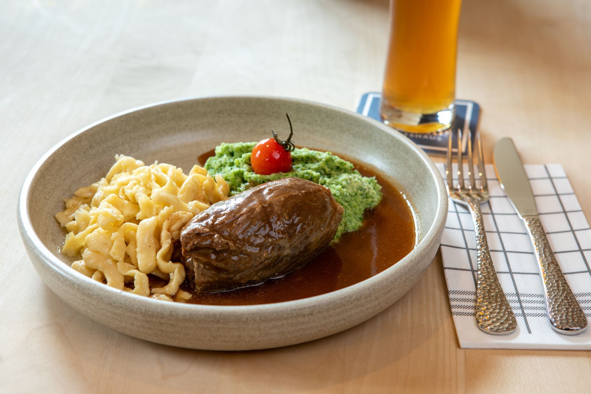 Rinderrolle mit Nudeln, grünem Püree und Soße in einer Schüssel, mit Bierglas und Besteck.